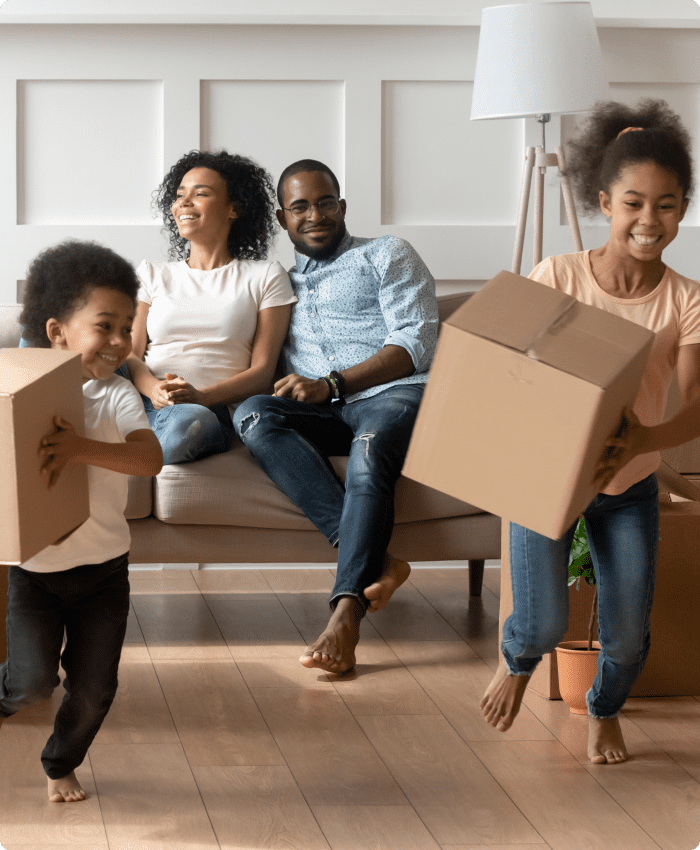 Imagem de familia, casal e duas filhas pequenas, na sala da casa, segurando caixas, demosntrando cada nova. Em mudança. A família são afro-descendentes. Página Sobre Nós.